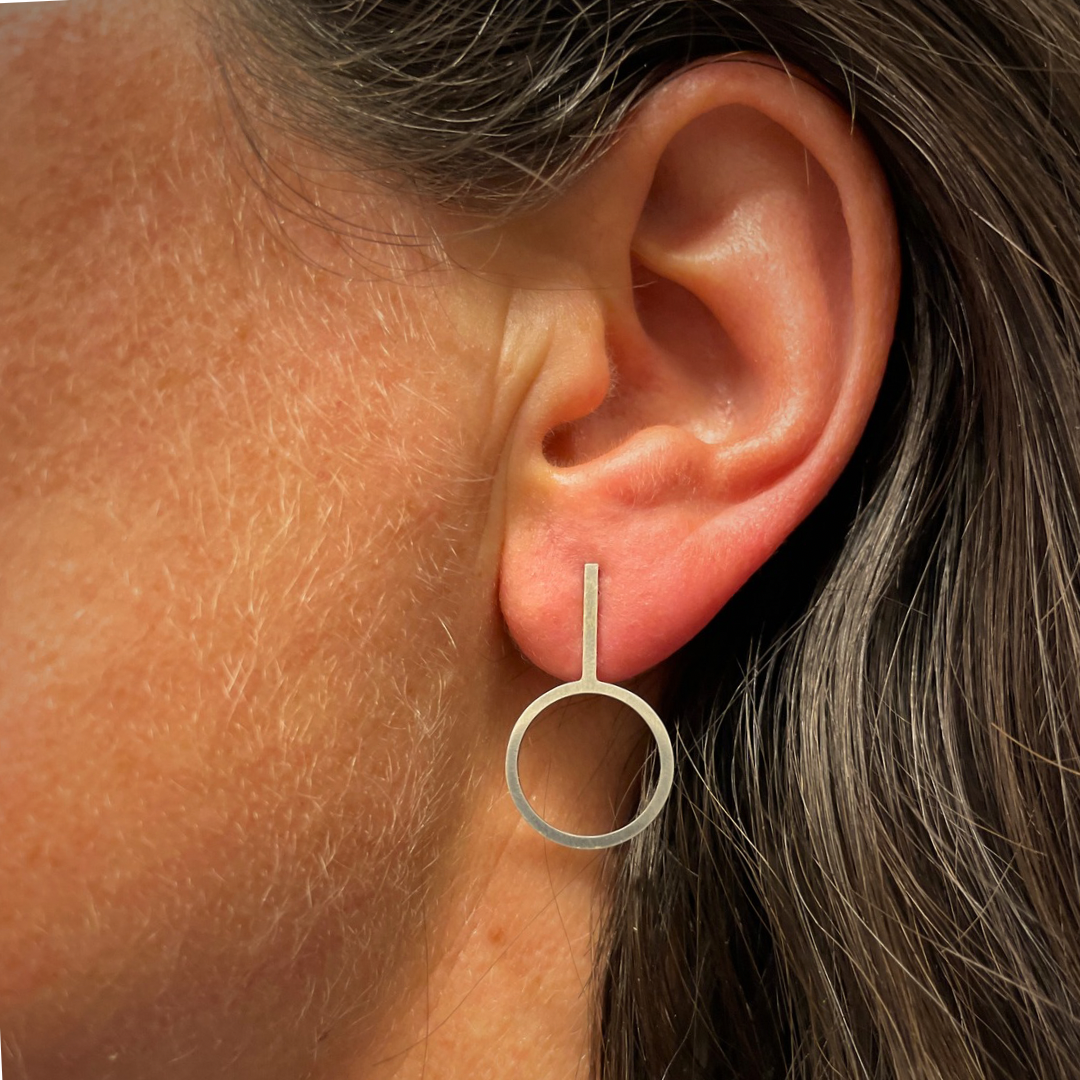 Close up of a woman’s ear who is wearing a circular sterling silver earring with a vertical wire attached to it with a total length of 2,4 cm.